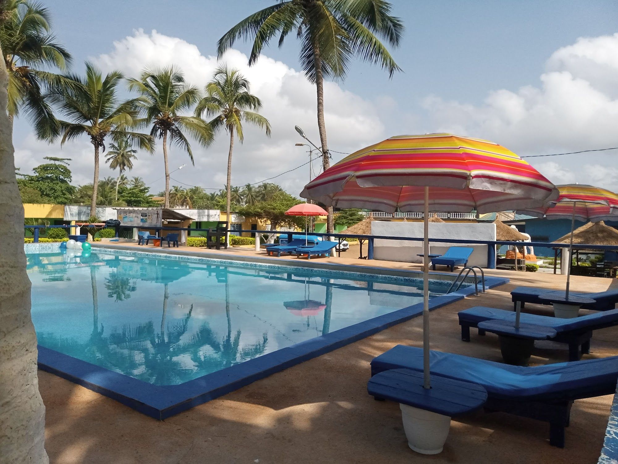 La piscine bordée de transats bleus et de cocotiers, un parasol rayé au premier plan